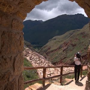 Una persona con una maglietta WeRoad si trova sotto un arco di pietra, ammirando le saline a terrazze in una valle di montagna.
