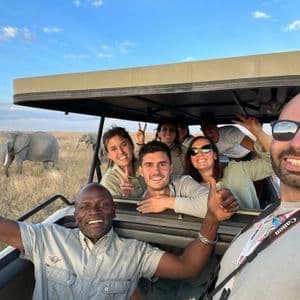 Un gruppo WeRoad in viaggio: persone sorridenti si scattano un selfie in un veicolo safari con elefanti sullo sfondo nella savana.