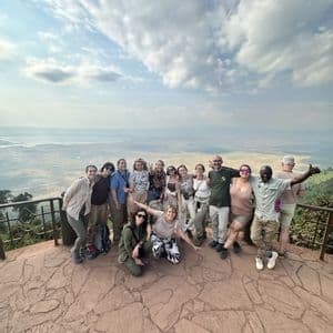 Un gruppo WeRoad in posa per una foto su un punto panoramico in pietra, con vista su una vasta valle e un lago sotto un cielo nuvoloso.