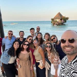 Un gruppo WeRoad si scatta un selfie sorridente su una spiaggia, con un ristorante nella roccia in mare sullo sfondo.