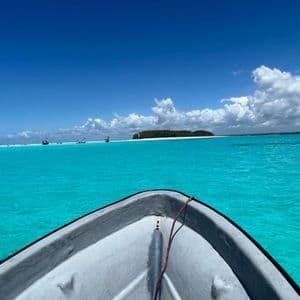 La prua di una barca naviga su acqua turchese verso un'isola lontana e alberata sotto un cielo blu con nuvole bianche.