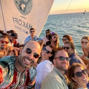 Un viaggio di gruppo WeRoad scatta un selfie sorridente insieme su una barca a vela in mare durante un tramonto dorato.