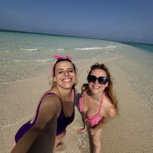 Due donne in costume sorridono per un selfie mentre sono in piedi in acqua limpida e bassa su un banco di sabbia.