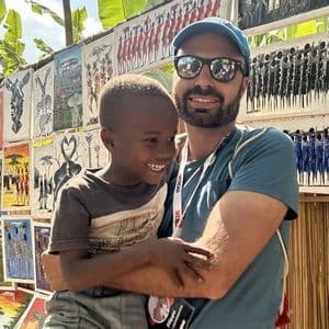 Un uomo di un viaggio di gruppo WeRoad sorride mentre tiene in braccio un bambino davanti a una bancarella d'arte all'aperto.