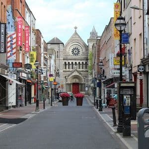 A view down a narrow city street lined with shops and signs, leading to a large stone church with a rose window at the end.