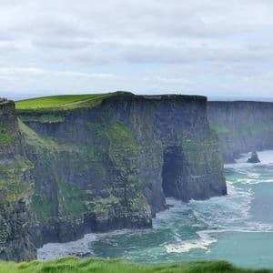 Una larga hilera de escarpados acantilados rocosos cubiertos de hierba verde vibrante se extiende a lo largo de la costa bajo un cielo nublado.