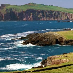 Des vagues blanches et écumeuses se brisent contre une côte escarpée, bordée de falaises verdoyantes, sous un ciel bleu clair.