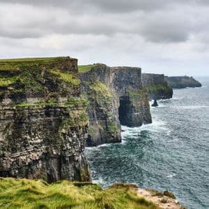 Des falaises escarpées et verdoyantes bordent le littoral, où elles rencontrent la mer agitée sous un ciel gris.