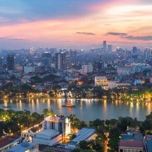 An aerial view of a sprawling cityscape with an illuminated lake at dusk, under a pink and blue sky.