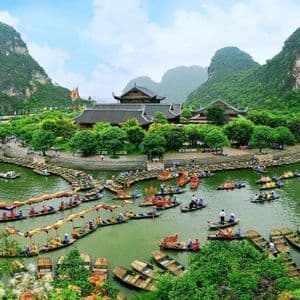 Dozens of traditional boats filled with people float on a river surrounding a temple complex at the base of lush, green mountains.