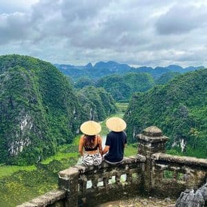 A man and a woman wearing conical hats sit on a stone wall, looking out over a lush green river valley surrounded by mountains.