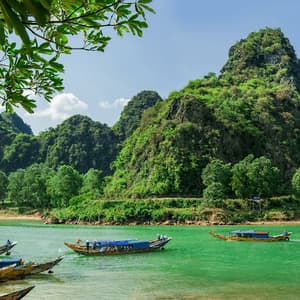 Traditional long boats on a vibrant green river at the base of lush, forest-covered mountains under a sunny sky.