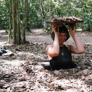 A smiling woman with glasses emerges from a hole covered with leaves in a forest, holding the leafy lid over her head.