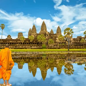 Two monks in orange robes stand by a lake, looking at an ancient stone temple reflected in the calm water.
