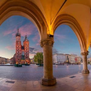Una iglesia histórica de ladrillo rojo con dos torres se erige en una plaza de la ciudad, observada desde debajo de arcos de piedra iluminados al atardecer.