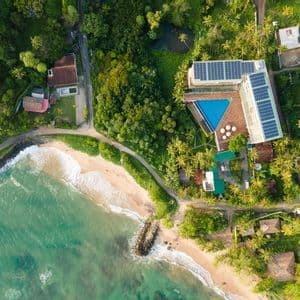 Vue aérienne d'un hôtel avec panneaux solaires et piscine triangulaire, situé à côté d'une plage de sable aux eaux turquoise.