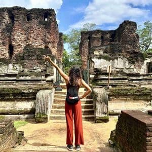 Une femme, sac à dos, vue de dos, le bras levé devant d'anciennes ruines de briques moussues.