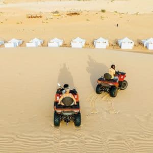 Vista aérea de dos personas de un viaje en grupo de WeRoad en quads por dunas de arena, con un campamento en el desierto visible al fondo.