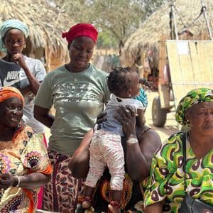 Un grupo de mujeres y niños con coloridos trajes tradicionales se encuentran en una aldea con chozas de techo de paja.