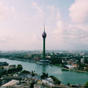 Ein hoher grüner Turm mit einer lotusförmigen Spitze steht inmitten einer Stadtlandschaft neben einem großen Gewässer.