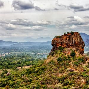 Un grand monolithe rocheux, brun-rougeâtre, s'élève au-dessus d'une vaste jungle verte et dense, avec des montagnes lointaines sous un ciel nuageux.