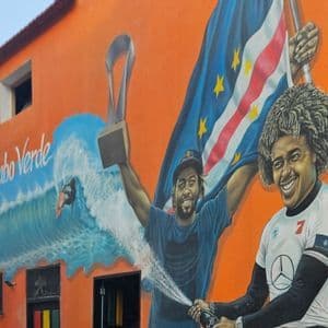 Ein farbenfrohes Wandgemälde an einem orangefarbenen Gebäude, das Sportler zeigt, die mit einer Trophäe und der Flagge von Cabo Verde feiern.