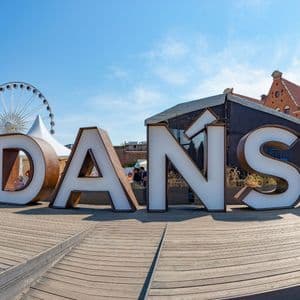 Ein großes, dreidimensionales Schild mit der Aufschrift 'GDAŃSK' auf einem Holzdeck, mit Riesenrad und Gebäuden im Hintergrund.