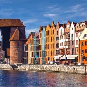 Eine Reihe farbenfroher Giebelhäuser und ein mittelalterlicher Backsteinkran säumen eine steinerne Uferpromenade und spiegeln sich unter blauem Himmel im Fluss.