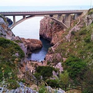 Un grande ponte ad arco attraversa una gola costiera rocciosa con un sentiero e scale che scendono verso una piccola insenatura sottostante.