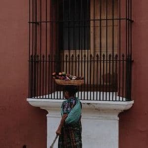 A woman in traditional clothing walks on a sidewalk, balancing a woven basket of produce on her head next to a reddish-brown wall.