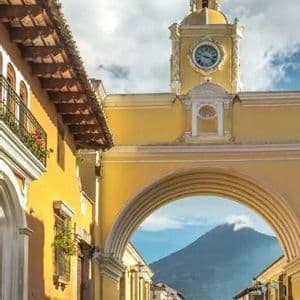 Un arco giallo con una torre dell'orologio si staglia su una strada di ciottoli, incorniciando la vista di un grande vulcano in lontananza sotto un cielo azzurro.