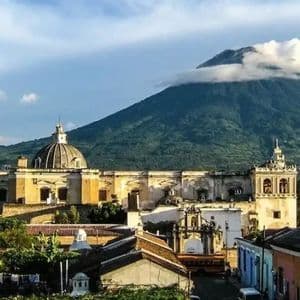 Una città coloniale con una grande chiesa a cupola si trova ai piedi di un vulcano verdeggiante, con la cima avvolta dalle nuvole, sotto un cielo azzurro.