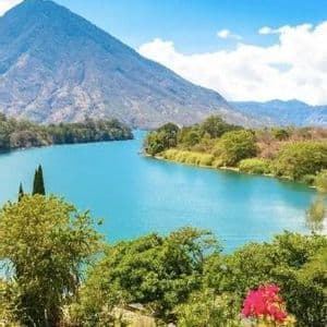 Un lago azzurro intenso si snoda attraverso una lussureggiante valle verde ai piedi di una grande montagna, con fiori rosa in primo piano.