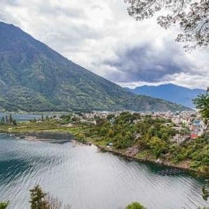 Una città lacustre si trova su una penisola ai piedi di una grande montagna boscosa sotto un cielo nuvoloso.