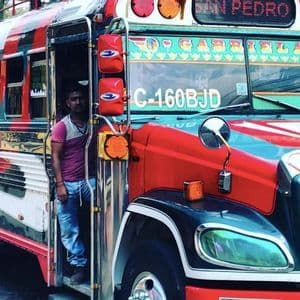 Un uomo con una maglietta rosa e jeans è in piedi sulla porta aperta di un autobus dai colori vivaci e con dettagli cromati.