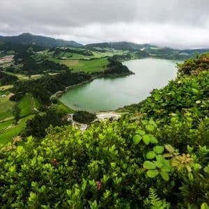 A high-angle view of a large green lake surrounded by rolling hills and patchwork fields, with lush foliage in the foreground.