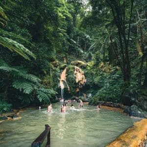 A WeRoad group trip swimming in a natural thermal pool with a waterfall, surrounded by a dense green forest.