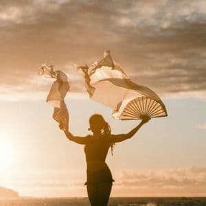 Une personne en silhouette devant un coucher de soleil se tient sur des rochers au bord de la mer, tenant deux grands éventails flottants.