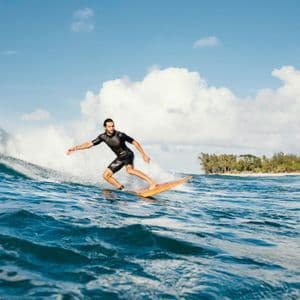 Un homme en combinaison de plongée noire surfe sur une vague bleue de l'océan, avec une plage de sable bordée d'arbres en arrière-plan.