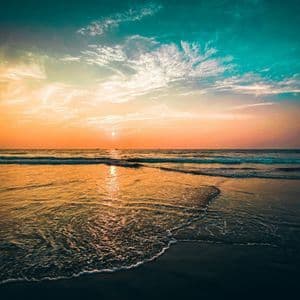 Le soleil se couche sur l'océan, projetant une lueur dorée sur l'eau tandis que les vagues déferlent sur une plage de sable sous un ciel turquoise.
