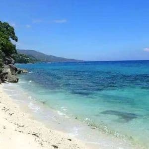 A white sand beach meets clear turquoise water next to a rocky, tree-covered shoreline under a blue sky.
