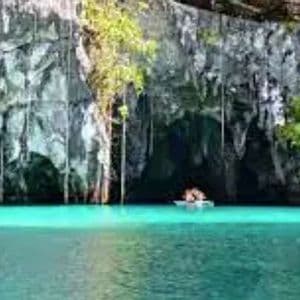 A small boat with people floats on turquoise water at the entrance of a large sea cave with rocky cliffs and hanging vines.
