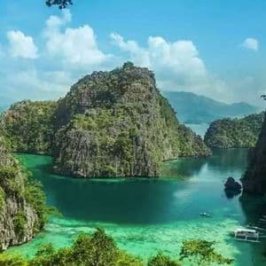A high-angle view of a tropical lagoon where lush, green-covered limestone cliffs rise from clear turquoise water under a partly cloudy sky.