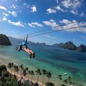 A person on a zip-line glides over a tropical beach with palm trees and turquoise water, with islands in the background.