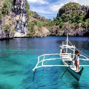 A person navigates a traditional outrigger boat across a turquoise lagoon, enclosed by steep, jungle-covered rock formations.