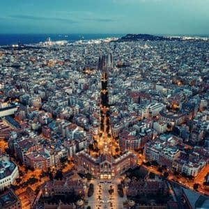 Une ville tentaculaire vue d'en haut au crépuscule, avec ses rues illuminées formant un quadrillage et l'océan au loin.