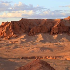 Des formations rocheuses rouges et des collines sablonneuses dans un vaste paysage désertique sont éclairées par le soleil de l'heure dorée sous un ciel partiellement nuageux.