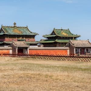 Un monastère mongol aux toits de tuiles vertes et au mur d'enceinte rouge, vu d'un champ d'herbe sèche sous un ciel bleu clair.