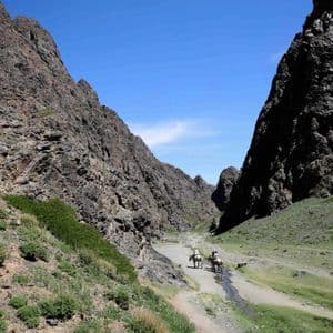 Deux personnes en voyage de groupe WeRoad montent à cheval à travers une vaste vallée montagneuse et rocheuse, près d'un ruisseau sous un ciel bleu.