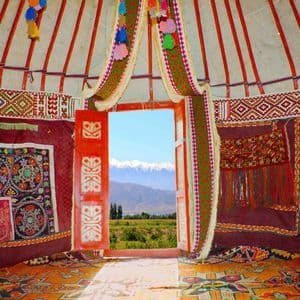 L'intérieur d'une yourte traditionnelle et ornée, avec une porte rouge ouverte encadrant une vue sur des montagnes enneigées.